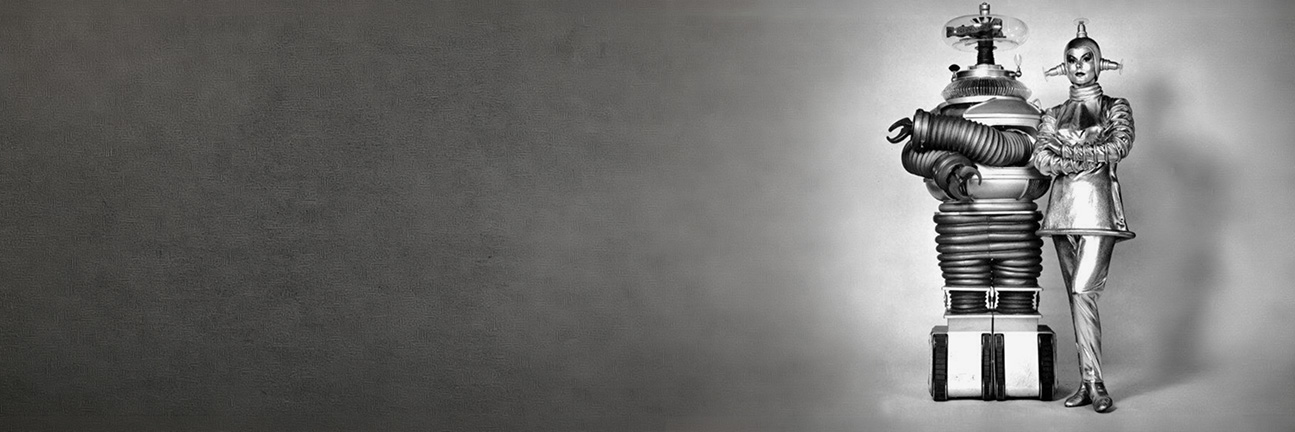 A classic television robot and a woman in a silver space-age costume standing at the right of a grey backdrop.