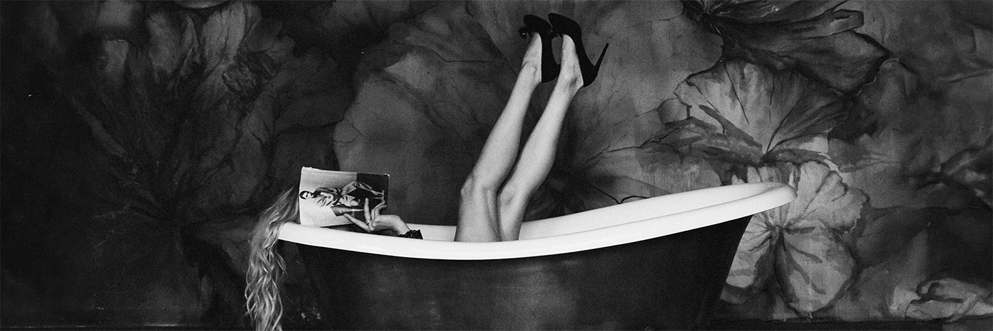 A woman reclining in a clawfoot bathtub reading a magazine, legs raised in black heels.