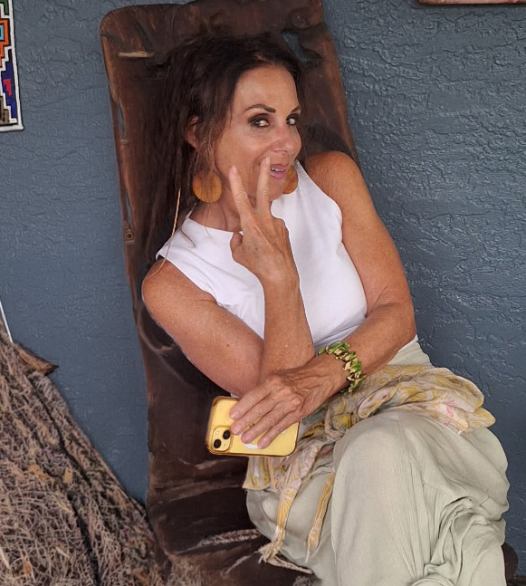 Caren Corso seated in a carved wooden chair, smiling and holding up two fingers in a peace sign.